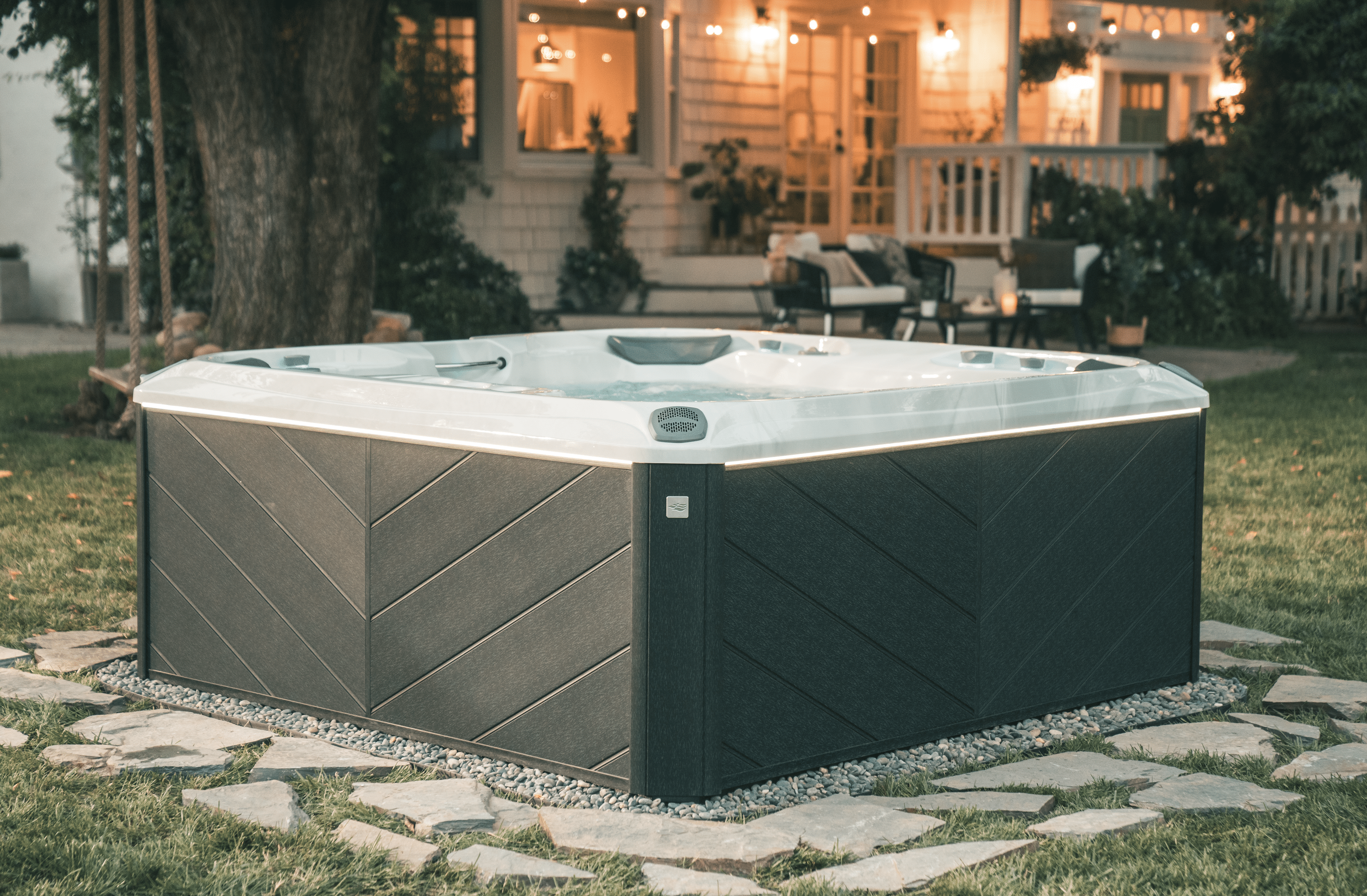 Hot tub in a backyard landscape in Bethel, CT. Stone patio underneath hot tub, illuminated back porch in background.