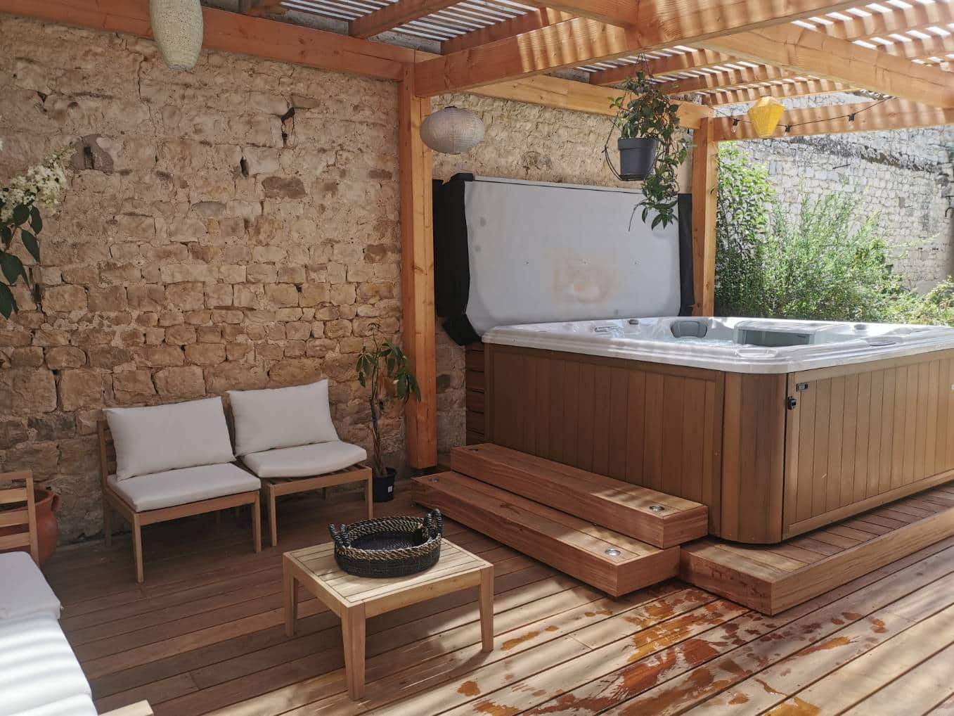 Brown sundance spa on a pergola-covered outdoor patio in Somers, NY. Hot tub uncovers with chairs and small table nearby, all atop wood planks and near exposed brick wall.
